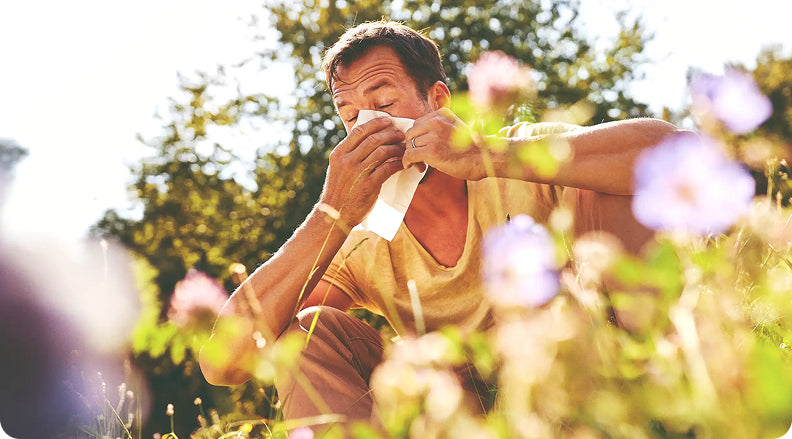 Uomo seduto in un campo fiorito mentre si soffia il naso, probabilmente a causa di un'allergia stagionale
