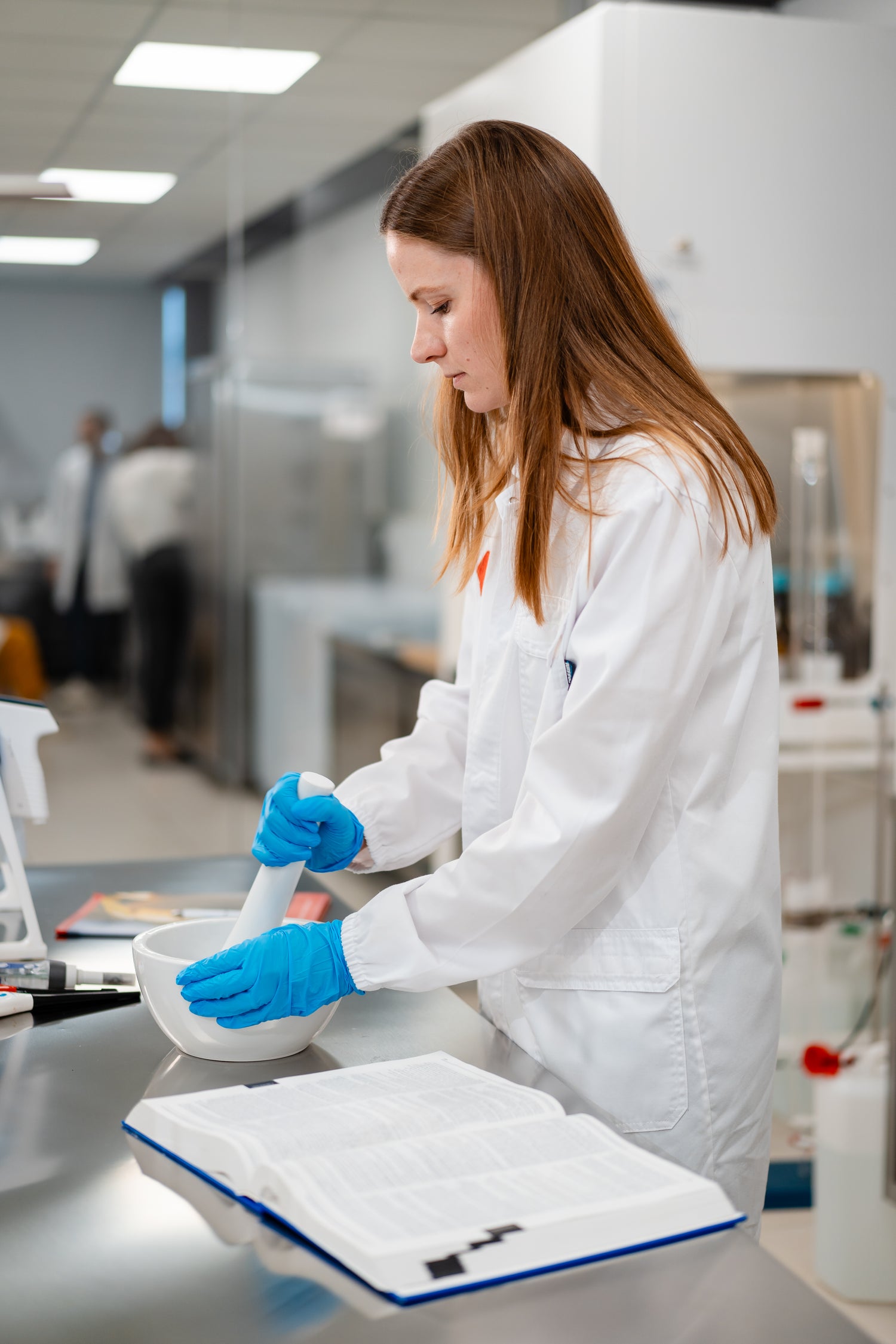 Ricercatrice in camice bianco e guanti blu che lavora in laboratorio consultando un manuale aperto