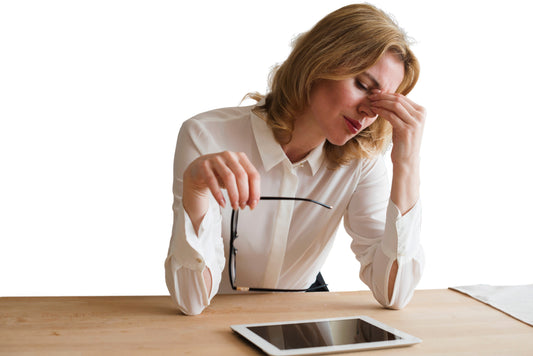 Donna in ufficio che manifesta affaticamento e stress durante il lavoro al computer, con occhiali in mano e tablet sulla scrivania.