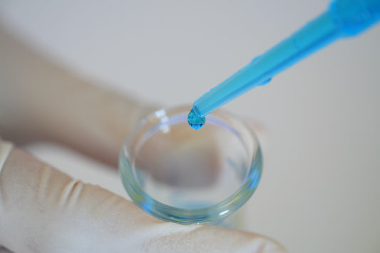 Pipetta con goccia di liquido blu sopra una capsula di Petri tenuta da una mano guantata in laboratorio.